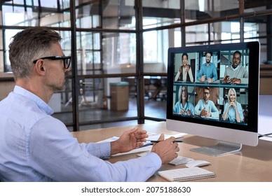 Caucasian businessman ceo in glasses having working videocall on financial report with multiethnic colleagues using personal computer in modern corporation global office. Over shoulder view.