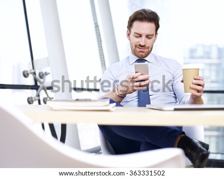 caucasian businessman sitting at desk looking at mobile phone holding cup of coffee in office.