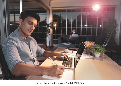 Caucasian Businessman Sitting Happily On Desk In Living Room Under Warm Lamp With Flare Cicle And Twilight Blue Sky Outside House. Idea For Working Late And Lifestyle Of Freelance, Working At Home.