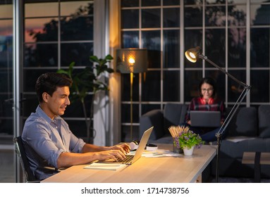 Caucasian businessman sitting happily on desk in living room under warm lamp and twilight blue sky outside house in background. Idea for working late and lifestyle of freelance, working at home couple - Powered by Shutterstock