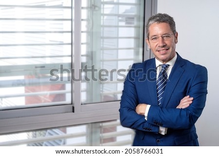 CAUCASIAN BUSINESSMAN STANDING NEXT TO A WINDOW AT THE OFFICE WITH FOLDED ARMS SMILING AND LOOKING AT CAMERA. CONCEPT OF EXECUTIVE, MANAGER, DIRECTOR OR ENTREPENEUR.