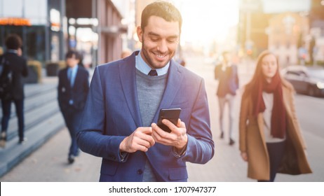 Caucasian Businessman in a Suit is Using a Smartphone on a Street in Downtown. Other Office People Walk Past. He Smiles and Looks Successful. He's Browsing Web on his Device. Shot with Warm Sun Flare. - Shutterstock ID 1707947377