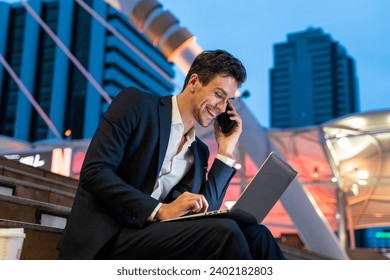 Caucasian businessman talk on mobile phone while work in city at night. Attractive handsome male employee worker using laptop computer to communicate internet chatting online to complete urgent job. - Powered by Shutterstock