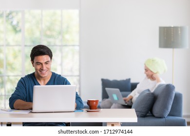 Caucasian Businessman Using Notebook Computer Working At Desk In Living Room While Woman With Towel Binding On Head Sitting And Holding Laptop In Blur Background. Idea For Calm And Happy Home Base Job