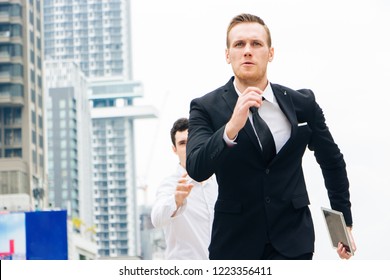 Caucasian Businessman Wear Black Suits And Tie In His Hand Holds The Tablet Running Forward With A Determined Expression And Assurance. Behind Him Was A Man Raising His Hands. Backdrop Is A Building.