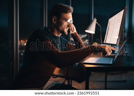A caucasian businessman works late at night in an office, discussing a project over the phone. He is focused and determined, balancing technology and communication for success.