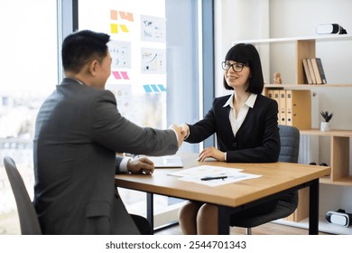 Caucasian businesswoman and Asian businessman shaking hands in modern office setting. Business professionals reaching agreement, sealing contract, partnership success symbolized. - Powered by Shutterstock