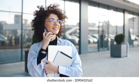Caucasian businesswoman with curly hair talking on phone and smile while holding a laptop. Urban lifestyle.