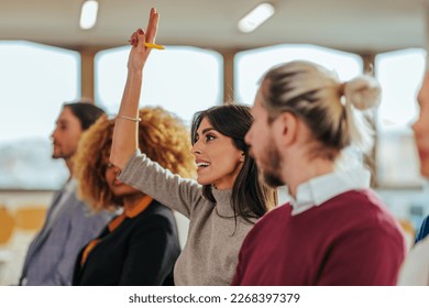 A Caucasian businesswoman is holding her hand up in a conference room to ask the speaker a question.
