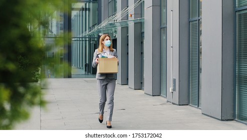 Caucasian businesswoman in medical mask walking outdoors with box of stuff leaving business. Female office worker lost her job. Unemployment rate growing at pandemic. Fired woman coming back home. - Powered by Shutterstock