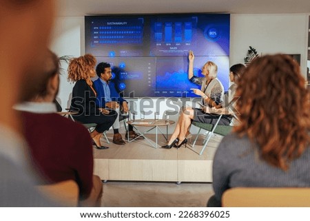 A Caucasian businesswoman is presenting a graph on a led display during a panel discussion about development in a corporate conference.