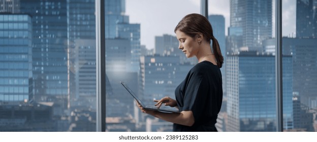 Caucasian Businesswoman Standing In Her Corner Office Next To Big Window With City View And Using Laptop Computer. Successful Business Manager Reviewing Tasks, Reading Emails. - Shutterstock ID 2389125283