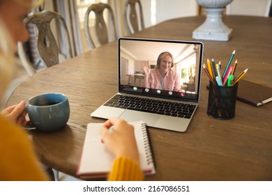 Caucasian businesswoman writing in notepad while video conferencing with teammate over laptop. coffee, unaltered, wireless technology, teamwork, meeting, discussion, business and work from home.