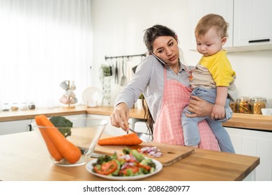 Caucasian Busy Mother Doing Housework With Baby Boy Toddler In Kitchen. Beautiful Single Mom Use Phone Call For Work And Cook Foods Prepare Breakfast For Little Kid Son In House. Family-housekeeping.