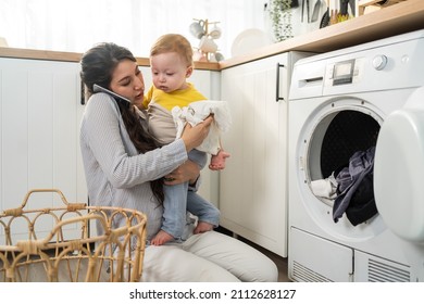 Caucasian busy mother doing housework with baby boy toddler in kitchen. Beautiful mom use phone call for work and put clothes to washing machine with her son play around in house. Family-housekeeping. - Shutterstock ID 2112628127