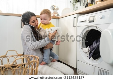 Caucasian busy mother doing housework with baby boy toddler in kitchen. Beautiful mom use phone call for work and put clothes to washing machine with her son play around in house. Family-housekeeping.