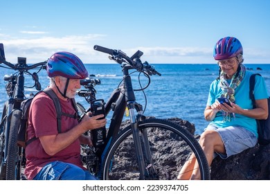 Caucasian carefree senior couple at the beach with electric bicycles in a sunny day. Man checks the battery while woman uses phone - Powered by Shutterstock