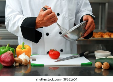 Caucasian chef mixing something in a bowl with fresh vegetables on a cutting board in front of him in a restaurant kitchen.