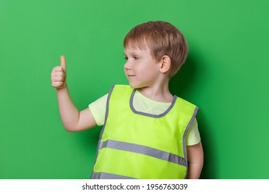 Caucasian Child Boy In Light Color Bright Vest With Reflective Stripes Shows Thumbs Up Gesture, Great, And Smile. Studio Shot On Green Background, Safety And Traffic Rules Concept.