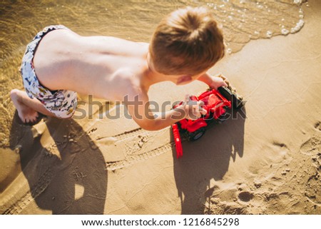 Caucasian child boy plays toy red tractor, excavator on sandy beach by the river in shorts at sunset day.