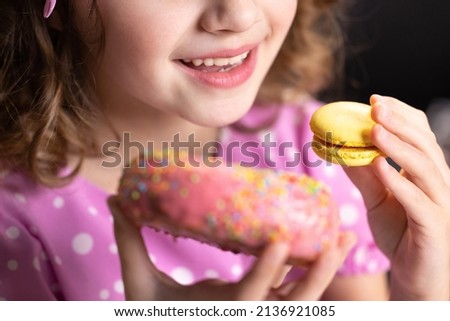 Caucasian child school girl eating donut and macaron at the same time. Adorable kid girl biting piece of donut macaron while sitting at table at home. Sweet addiction concept