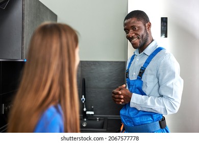 Caucasian Client Woman Is Grateful To Smiling Afro Plumber For Repair Glitch In Kitchen, Confident Handyman In Blue Overalls Having Talk With Female Client, Discussing Repair. Focus On Plumber