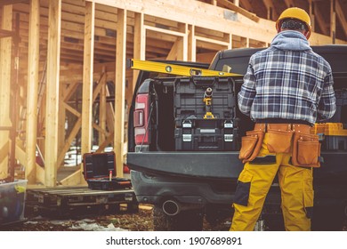 Caucasian Construction Contractor In His 40s And His Modern Black Pickup Truck. Men Preparing His Tools For The Job. Wooden House Skeleton In Background. 