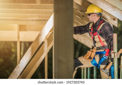 Caucasian Construction Contractor In His 40s Wearing Safety Harness And Hard Hat. Wooden House Skeleton Frame Building. Industrial Theme.