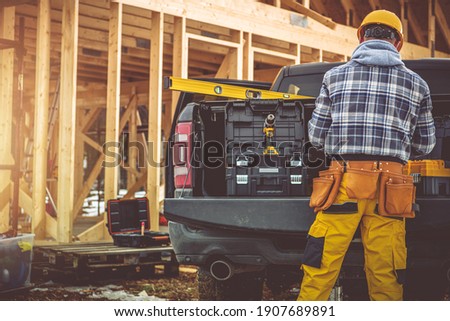 Caucasian Construction Contractor in His 40s and His Modern Black Pickup Truck. Men Preparing His Tools For the Job. Wooden House Skeleton in Background. 