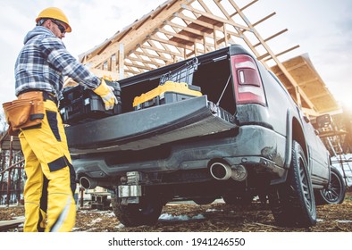 Caucasian Construction Contractor Worker In His 40s Taking Tools Boxes From Bed Section Of His Pickup Truck. Newly Constructed Building In Background.