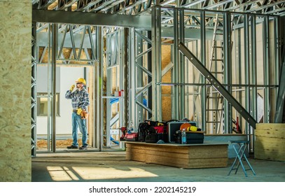 Caucasian Construction Worker Standing On Building Site Surrounded By Steel House Frame With Wooden Elements. Wearing Yellow Safety Hard Hat And Communicating With Other Workers Via Walkie Talkie.