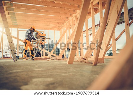 Caucasian Contractor Carpenter Worker in His 40s Using Commercial Grade Circular Saw in Construction Zone. Industrial Theme. Wooden Skeleton Framing Building.