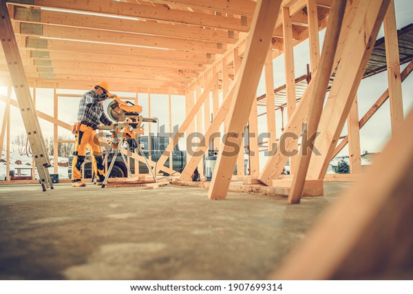 Caucasian Contractor Carpenter\
Worker in His 40s Using Commercial Grade Circular Saw in\
Construction Zone. Industrial Theme. Wooden Skeleton Framing\
Building.