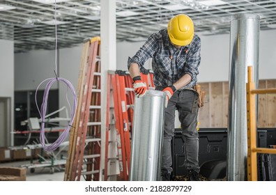 Caucasian Contractor In Yellow Hard Hat Moving The Ventilation Pipe During Installation Of HVAC Temperature Control System In The Commercial Warehouse Hall. Construction Site In The Background.