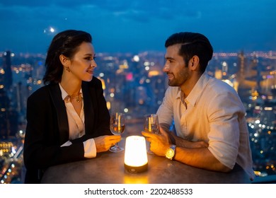 Caucasian Couple Celebrating At Luxury Skyscraper Rooftop Bar At Night. Confident Man And Woman Couple Enjoy City Lifestyle Nightlife Having Dinner And Cocktail Drink At Outdoor Restaurant In Evening.