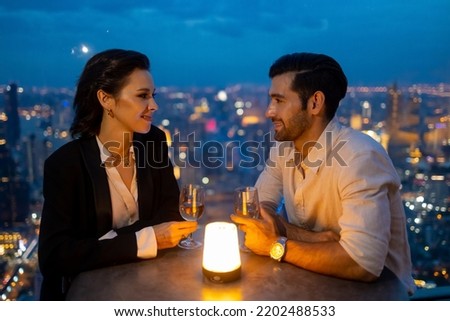 Caucasian couple celebrating at luxury skyscraper rooftop bar at night. Confident man and woman couple enjoy city lifestyle nightlife having dinner and cocktail drink at outdoor restaurant in evening.