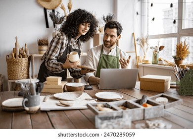 Caucasian couple of co-workers work on small pottery production business. Mature man and young curly-haired woman in aprons use laptop to take inventory of products on store website. - Powered by Shutterstock