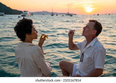 Caucasian Couple Enjoy Outdoor Luxury Party Drinking Champagne With Talking Together While Catamaran Boat Sailing At Sunset. Man And Woman Relax With Outdoor Lifestyle On Summer Travel Vacation