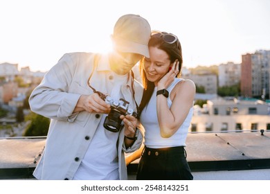 Caucasian couple enjoys reviewing photos together on sunny rooftop with cityscape backdrop. Man shows woman camera screen, memories at sunset. Warm light, connection and love. Casual urban background. - Powered by Shutterstock