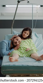 Caucasian Couple Expecting Child In Hospital Ward While African American Nurse Preparing IV Drip Bag For Pregnant Woman At Maternity Clinic. Husband And Wife Waiting For Baby Delivery