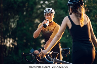 Caucasian couple practicing while riding a bike outside and taking a break from it to rest. Focus on a man sitting on his bike and drinking water. Concept of healthy lifestyle and outdoor sport. - Powered by Shutterstock