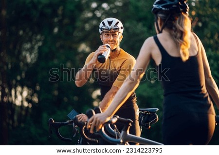 Caucasian couple practicing while riding a bike outside and taking a break from it to rest. Focus on a man sitting on his bike and drinking water. Concept of healthy lifestyle and outdoor sport.