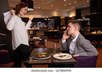 Caucasian Couple Quarreling In Restaurant. Woman Yell At Man Talking On Phone. Lady Stand In A Fury, Want To Go Away From Him