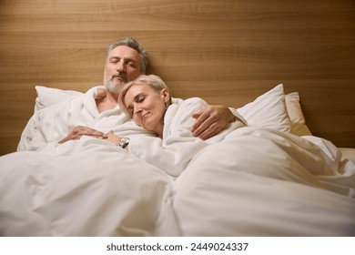 Caucasian couple wearing bathrobes embracing and sleeping on bed in hotel room - Powered by Shutterstock