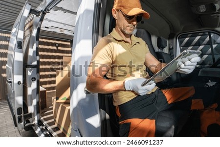 Caucasian courier seating inside delivery van and performing parcel documentation on his tablet.