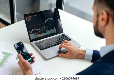 Caucasian crypto trader investor using cellphone and laptop for cryptocurrency financial market analysis, buying or selling cryptocurrency.Close-up of laptop and smart phone screen with stock diagrams