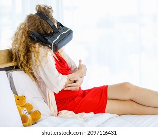 Caucasian Curly Long Hair Pregnancy Female Mother In Red Maternity Dress Pajama With Rope Wears Vr Goggles Sitting Smiling Lay Down On Bed Learning Study Practice Virtual Reality Babysitting Online.