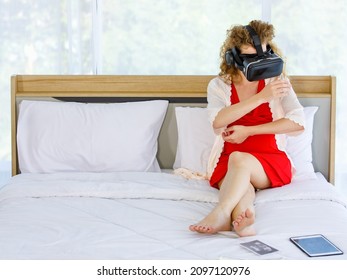 Caucasian Curly Long Hair Pregnancy Female Mother In Red Maternity Dress Pajama With Rope Wears Vr Goggles Sitting Smiling Lay Down On Bed Learning Study Practice Virtual Reality Babysitting Online.