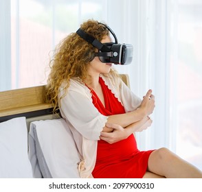 Caucasian Curly Long Hair Pregnancy Female Mother In Red Maternity Dress Pajama With Rope Wears Vr Goggles Sitting Smiling Lay Down On Bed Learning Study Practice Virtual Reality Babysitting Online.