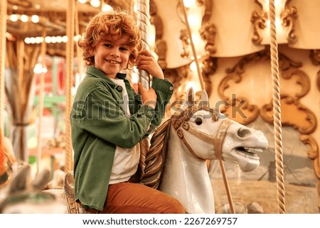 Caucasian curly-haired blonde boy ride a vintage horse carousel in the evening at an amusement park on a weekend.